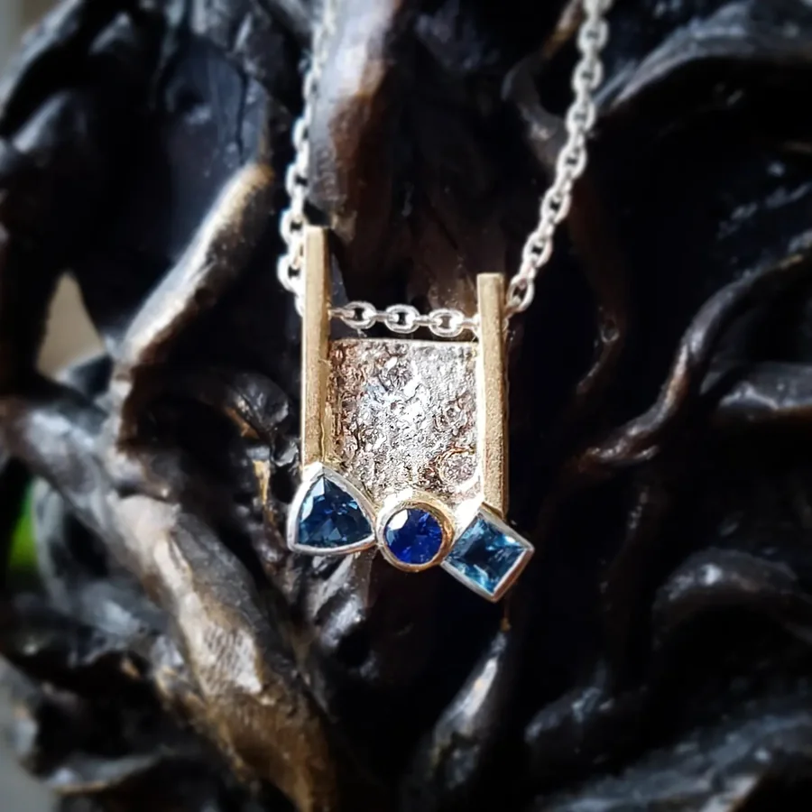 A textured silver pendant flanked by two gold posts and set with three blue stones, against dark driftwood.