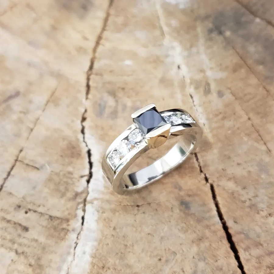 A white gold ring set with a princess cut black diamond and channel set white diamonds, on weathered pale wood.