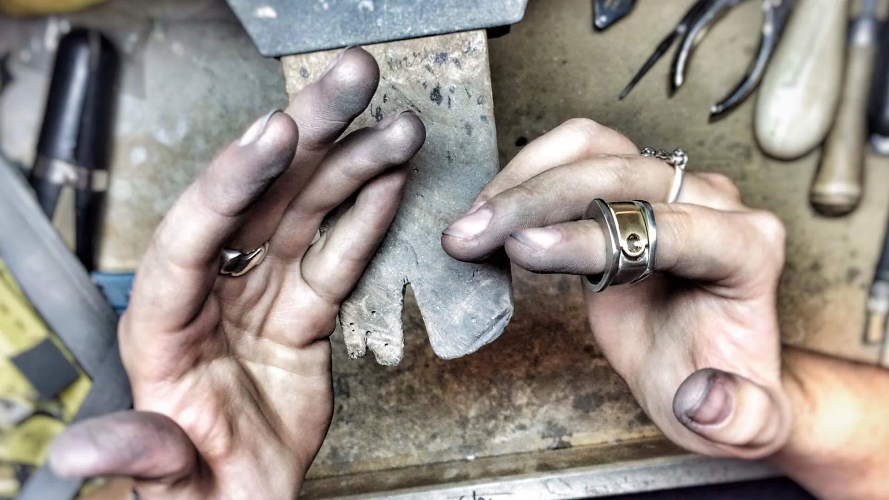Kat's hands at the workbench, holding a metal template against a bench pin, with pliers and burnishers laid out around them.