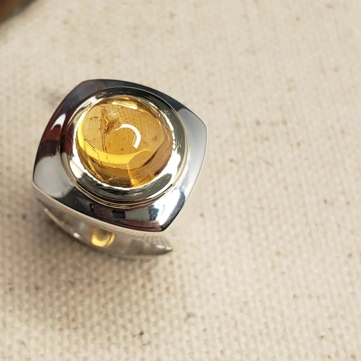 A wide silver ring holding a domed golden citrine cabochon in a gold bezel, on cream woven fabric.