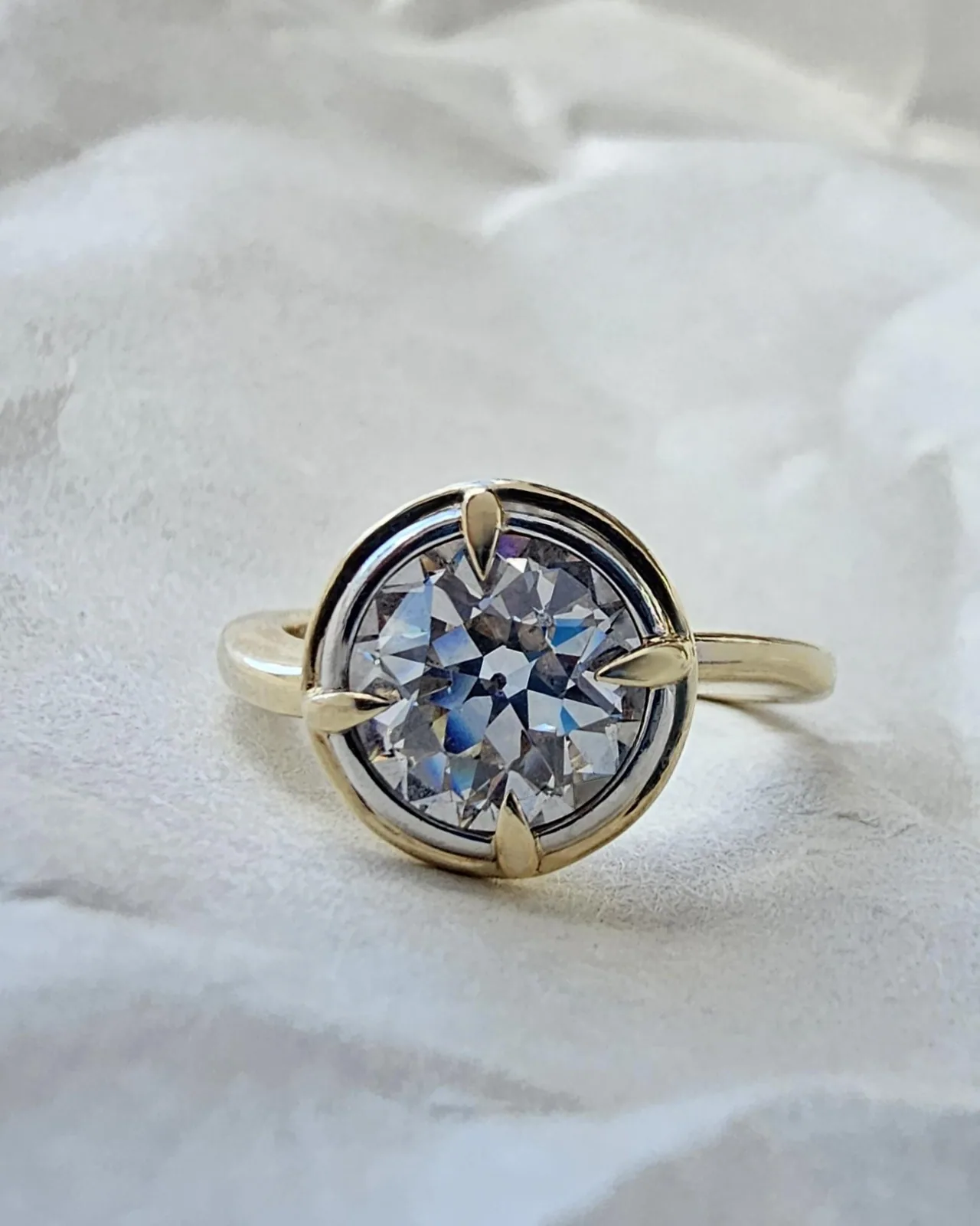 A close view of a round old cut diamond held by four gold claws in a compass style bezel on a plain gold band, against soft white paper.