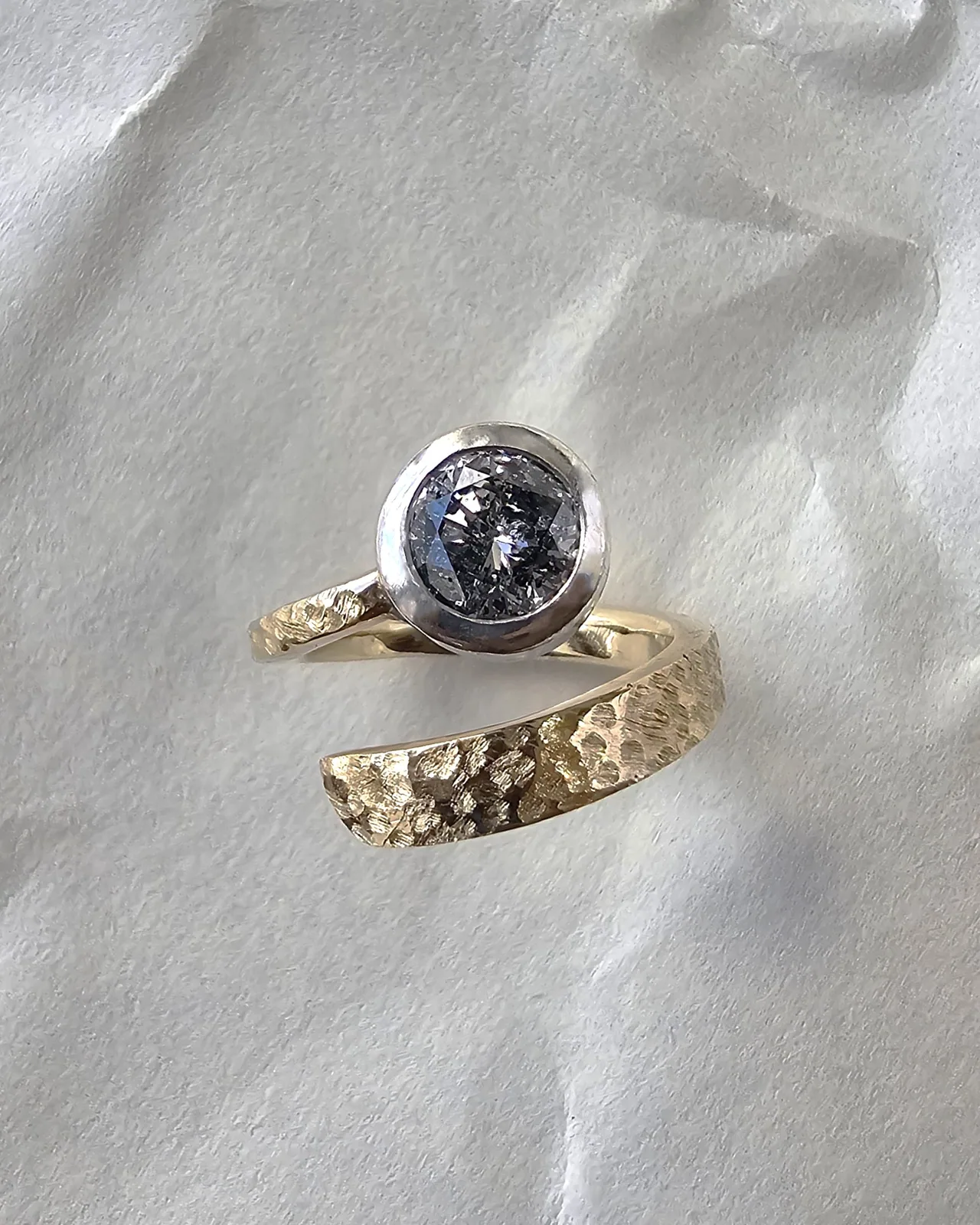 A round salt and pepper diamond bezel set in white metal on a hammered gold bypass band, photographed from above on crumpled white paper.