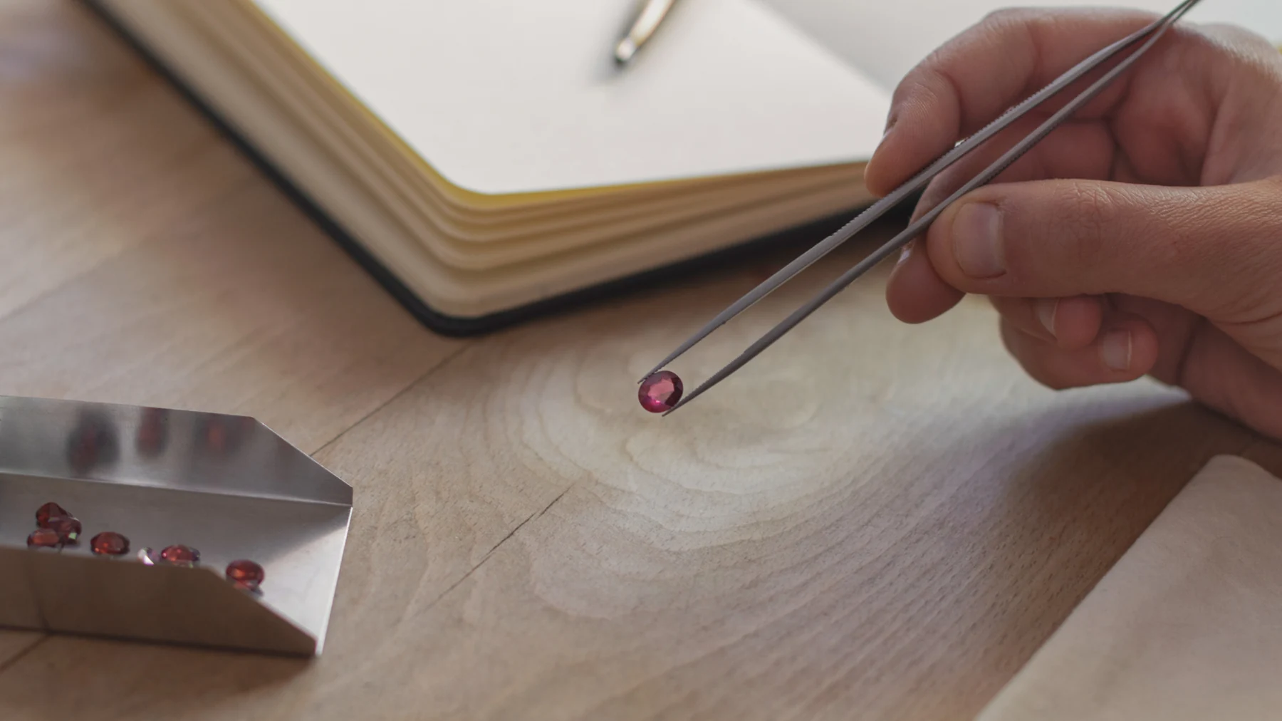 A hand holding tweezers with a rhodolite garnet above a metal parcel tray of loose red stones.