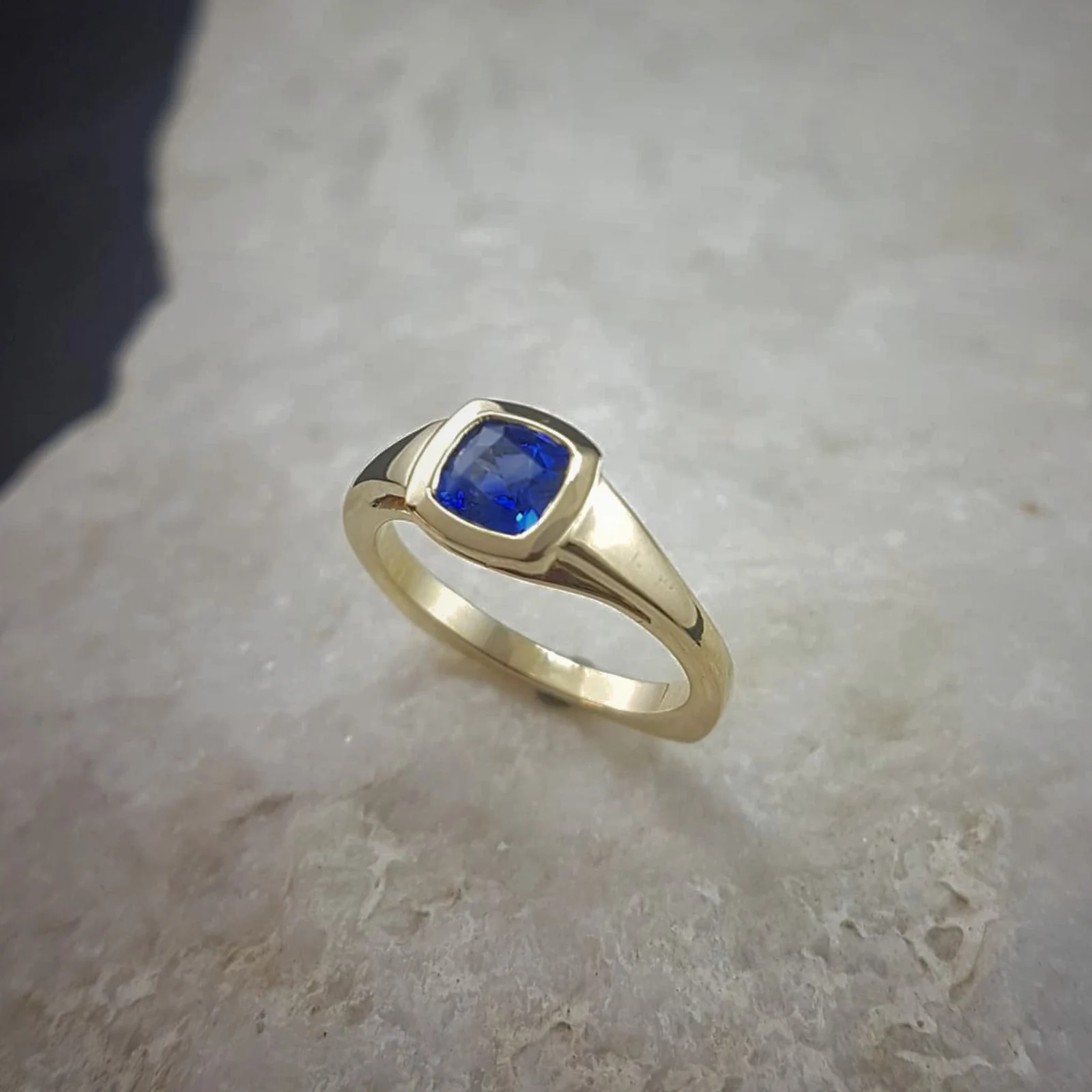 The finished ring: a deep blue cushion cut sapphire bezel set in 18K yellow gold, photographed on white stone.