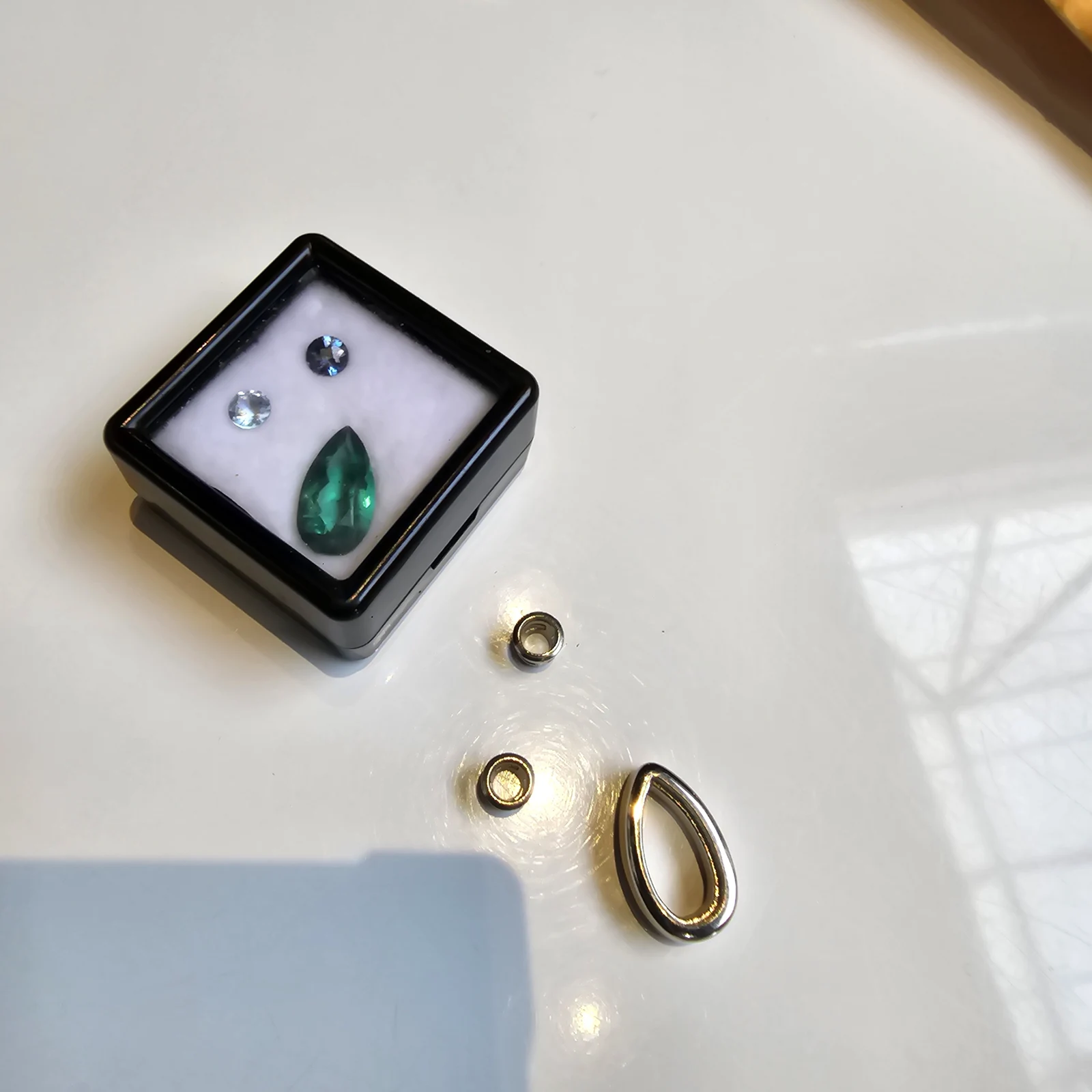 Loose stones in a gem case beside gold components: a pear cut emerald, a round blue sapphire, a pale round stone, two gold bezel cups and a pear shaped gold setting, on a white surface in window light.